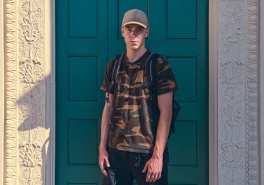 Yeşil bir kapının önünde, üzerinde kamuflaj tişörtü olan genç bir adamın portresi.