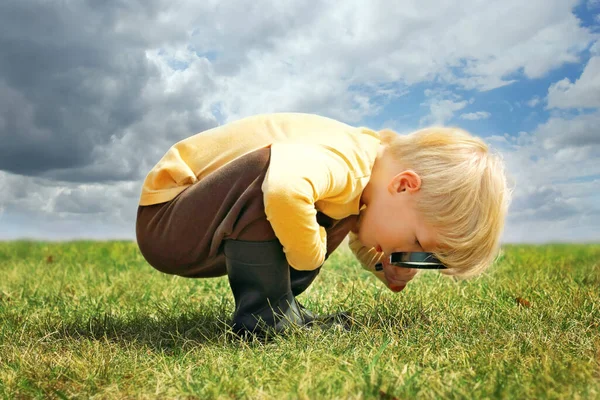 A little boy is exploring nature outside by looking at the grass through a magnifying glass. 