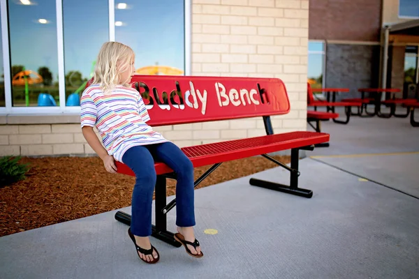 一个寂寞的小女孩正坐在学校的长椅上等着一个朋友和她一起在操场上玩耍 — 图库照片