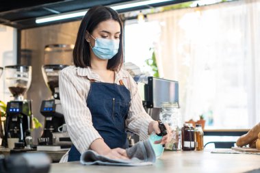 Kafe garsonu maske takıyor, mağazayı açmak için dezenfektanlı temiz bir restoran. Asyalı çekici genç bir kadın, Covid19 kilitlendikten sonra dükkanı açmadan önce restoranda dezenfekte, silme ve ovalama yapıyor..
