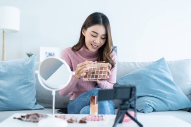 Evdeki kozmetiği incelemek için kamerayla Asyalı kadın video blogcu konuşması. Güzel blogcu Nüfuzlu Kız kanepeye oturur ve canlı yayında izleyicilere makyaj malzemelerini gösterir..