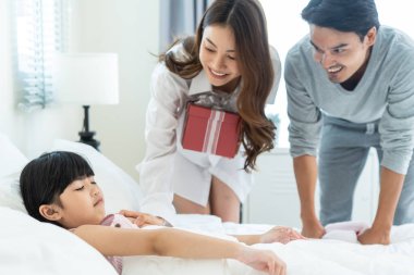 Asyalı mutlu aile, genç anne ve babalar sürpriz bir şekilde kızı hediyeyle uyutur. Çekici erkek ve kadın çift yataktaki küçük çocuklarını uyandırıp doğum günü hediyesi vermek ve geleneksel partiyi kutlamak için