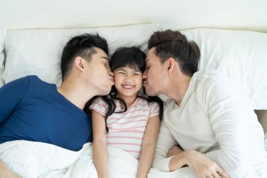 Yakışıklı eşcinsel bir ailenin portresi ve küçük bir kız çocuğu yatak odasında. Çekici romantik erkek lgbt çifti yatağa uzanıp küçük sevimli kızı öpüyor, evde ebeveynliğin tadını çıkarıyor..