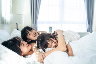 Asyalı mutlu çocuklar sabah annelerini uyandırmak için yatağa atlıyorlar. Küçük kız kardeş kızları, sevgi dolu anneleriyle birlikte mutlu mesut oyun oynarlar. Etkinlik ilişkisi kavramı