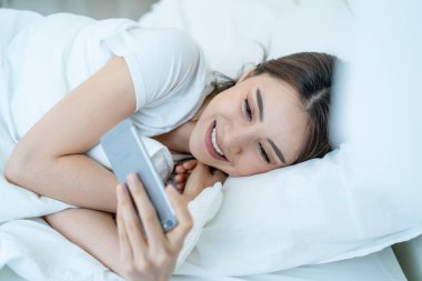 Asyalı çekici kadın sabah evde cep telefonu sohbetini kullanıyor. Mutlu, sıradan, güzel bir kadın yatak odasında uzanıyor. Tatil haftasonunun tadını çıkarın. Evdeki teknolojiyle akıllı telefon iletişimi kurun..