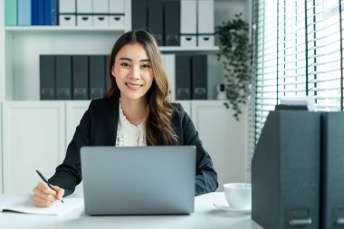 Ofiste çalışırken gülümseyen Asyalı güzel iş kadını portresi. Resmi kıyafetli çekici profesyonel kadın çalışan iş yerinde masaya oturur, dizüstü bilgisayar kullanır ve kameraya bakar..