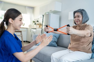 Asyalı engelli yaşlı kadın hemşirenin desteğiyle fizyoterapiste gidiyor. Kıdemli engelli kadın oturma odasında kanepede oturur evde bakım gören hastalar için direnç bandı egzersizi yapar.