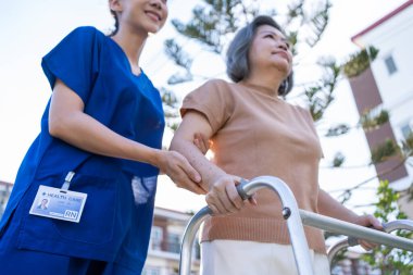 Asyalı bakıcı hemşire dışarıda yürüyen yaşlı engelli kadına destek oluyor. Güzel kız doktor yaşlı, olgun, engelli bir hastaya bakıcılık ve bakım evinde fizik tedavi uyguluyor..