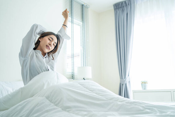 Азиатская красивая девушка в пижаме просыпается утром со счастьем. Привлекательная молодая женщина улыбается, чувствовать себя счастливым и расслабиться, а затем растяжения тела после вставания от сна на кровати в спальне на дому.