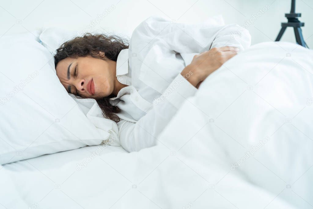 Latino beautiful sick girl in pajamas getting up from sleep in bedroom. Attractive young woman feel bad and painful after wake up on bed, having stomach ache or periods then clasping belly in morning.