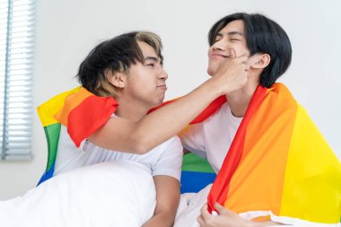 Asyalı yakışıklı bir adamın portresi gay bir ailenin LGBT bayrağı tutması ve gülümsemesi. Çekici romantik erkek lgbt çifti sabahları yatakta oturur ve gay gururu ve gökkuşağı arka planıyla kameraya bakarlar.