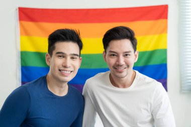Asyalı yakışıklı bir adamın portresi gay bir ailenin LGBT bayrağı tutması ve gülümsemesi. Çekici romantik erkek lgbt çifti sabahları yatakta oturur ve gay gururu ve gökkuşağı arka planıyla kameraya bakarlar.