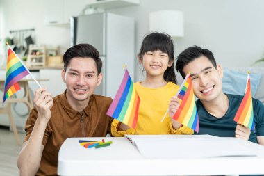 Asyalı LGBTQ erkek eşcinsel aile ve kız çocuğunun oturma odasında portresi. Çekici, yakışıklı erkek çift LGBTQ gökkuşağı bayrağı tutan ve kameraya bakan küçük tatlı çocukla ilgilenip vakit geçiriyor..