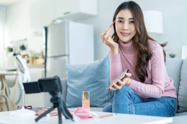 Evdeki kozmetiği incelemek için kamerayla Asyalı kadın video blogcu konuşması. Güzel Güzellik blogcusu Nüfuzlu Kız kanepeye otur ve canlı yayın yap, vakıf şovu yap, evdeki takipçilere makyaj yap.