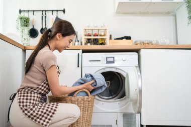 Asyalı güzel kadın evdeki çamaşır makinesine kirli çamaşır koydu. Çekici bir kız önlük giyer, evde çamaşır makinesine çamaşır doldurduğu için mutlu olur. Ev idaresi kavramı.