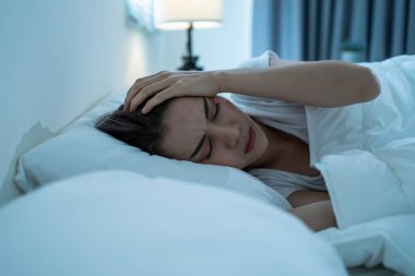 Asian beautiful upset depressed sick girl lying down on bed in bedroom. Attractive unhappy young woman in pajamas feel sad lonely and upset with headache and cannot sleep in dark night room at home.