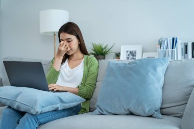Yorgun Asyalı iş kadını dizüstü bilgisayar kullanırken gözünü yaraladı. Genç ofis çalışanı kanepeye oturur aşırı çalışmaktan ve masajdan yorgun ve yorgun hisseder evde gözü kurur..