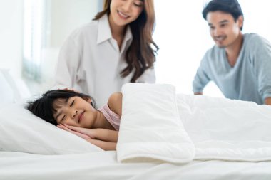 Asian loving parents take care of sleeping daughter in bedroom at home. Attractive happy family couple, mother and father puts blanket on comfortable asleep napping little baby kid on bed in morning.