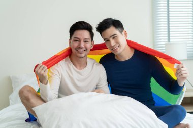 Asyalı yakışıklı bir adamın portresi gay bir ailenin LGBT bayrağı tutması ve gülümsemesi. Çekici romantik erkek lgbt çifti sabahları yatakta oturur ve gay gururu ve gökkuşağı arka planıyla kameraya bakarlar.