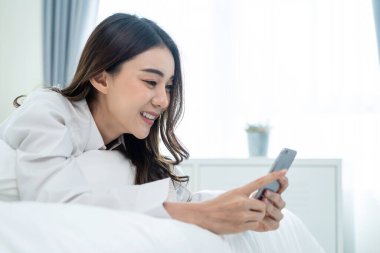 Asyalı çekici kadın sabah evde cep telefonu sohbetini kullanıyor. Mutlu, sıradan, güzel bir kadın yatak odasında uzanıyor. Tatil haftasonunun tadını çıkarın. Evdeki teknolojiyle akıllı telefon iletişimi kurun..