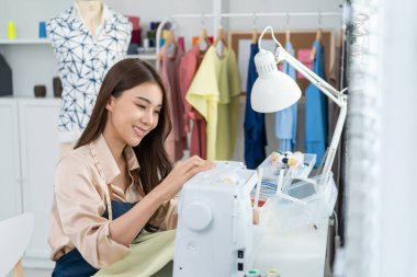 Asian beautiful tailor woman working on clothes in tailoring atelier. Attractive young female fashion designer pushes fabric material through a sewing machine making new handmade suit in workshop room