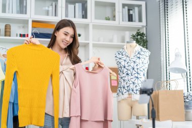 Asian beautiful woman sell cloth product online live streaming at home. Young female use mobile phone video call shows goods to customer and present detail. Remote buying and purchase shopping concept