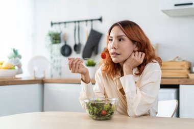 Asian young happy attractive woman eat green salad in kitchen at home. Beautiful girl feel joyful and enjoy eating vegetables healthy foods to diet and lose weight for health care wellness in house.