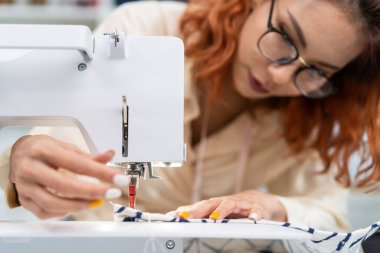 Asian beautiful tailor woman working on clothes in tailoring atelier. Attractive young female fashion designer pushes fabric material through a sewing machine making new handmade suit in workshop room