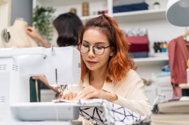 Asian beautiful tailor woman working on clothes in tailoring atelier. Attractive young female fashion designer pushes fabric material through a sewing machine making new handmade suit in workshop room