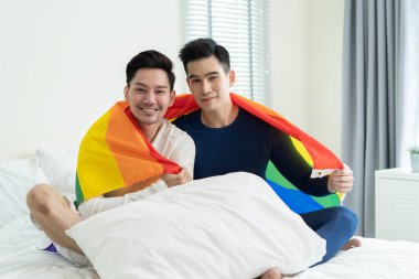 Portrait of Asian handsome man gay family holding LGBT flag and smile. Attractive romantic male lgbt couple sit on bed in bedroom in morning, look at each other with gay pride and rainbow background.