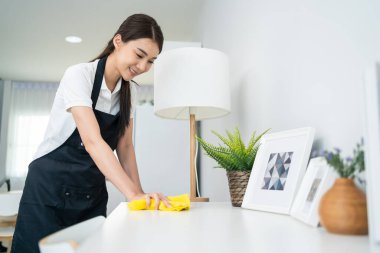 Asian cleaning service woman worker cleaning in living room at home. Beautiful girl housewife housekeeper cleaner feel happy and wiping messy dirty working table for housekeeping housework or chores.