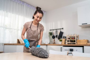 Asian cleaning service woman worker cleaning in kitchen room at home. Beautiful young housekeeper cleaner wear apron, and using feather duster wiping dirty table for housekeeping housework or chores.