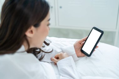 Asyalı bir kadın evdeki beyaz ekranla çevrimiçi video görüşmesinde konuşuyor. Pijamalı çekici bir kız, sabah yatak odasında uyandıktan sonra sanal bir uzaktan kumandada cep telefonuyla konuşmayı sever..