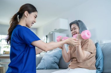 Asian Disabled old woman doing physiotherapist with support from nurse. Senior elderly handicapped female sit on sofa in living room using dumbbells workout exercise for patient in home nursing care.