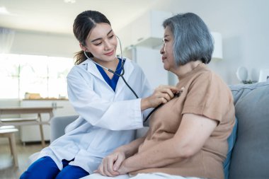 Huzurevindeki Asyalı hemşire engelli yaşlı kadınla ilgileniyor. Güzel terapist doktor kalp atışlarını, kanepedeki yaşlı kadın hastada steteskopla ölçer. Sağlık sigortası konsepti.