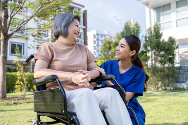 Asyalı bakıcı hemşire dışarıda tekerlekli sandalyeli yaşlı kadını destekliyor. Güzel doktor kız. Yaşlı, olgun, engelli bir hastaya yardım et. Bakımevinde fizik tedavi uyguluyor..