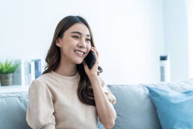 Asyalı güzel kadın kanepede oturur ve cep telefonuyla konuşur. Mutlu çekici genç kız evde vakit geçirir, rahatlar, iletişim kurar ve oturma odasında akıllı telefondan tartışır..