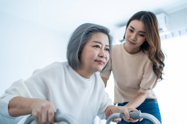 Asyalı genç kız, yaşlı engelli kadının evde Walker 'la yürümesini destekliyor. Güzel bir kız, yaşlı, olgun, engelli bir anneye yardım eder ve onunla ilgilenir.