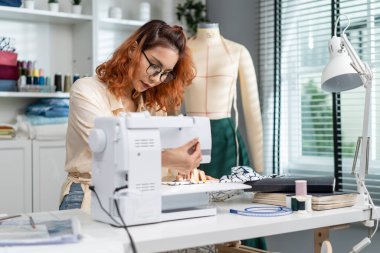 Asian beautiful tailor woman working on clothes in tailoring atelier. Attractive young female fashion designer pushes fabric material through a sewing machine making new handmade suit in workshop room