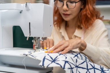 Asian beautiful tailor woman working on clothes in tailoring atelier. Attractive young female fashion designer pushes fabric material through a sewing machine making new handmade suit in workshop room