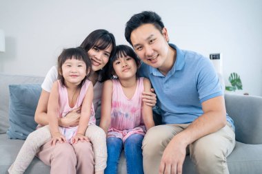 Portrait of Asian happy family spend time with kid together in house. Young little cute girl daughters and parents and sit on sofa smiling, look at camera in living room. Activity relationship concept