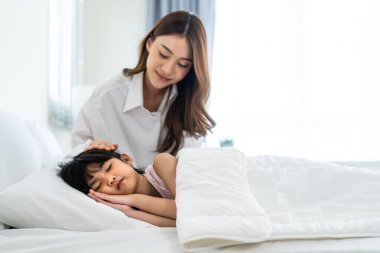 Asian loving parents take care of sleeping daughter in bedroom at home. Attractive happy family, beautiful mother puts blanket on comfortable asleep napping little baby kid on bed in morning in house.