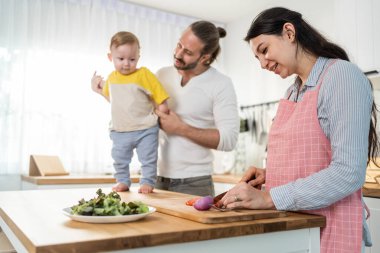 Caucasian beautiful parents cook food with baby boy toddler in kitchen. Happy family, Attractive young mother making healthy salad for lunch while husband father holding little kid son infant in house