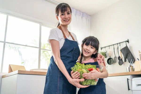 Asyalı güzel annenin portresi salata kasesi büfesini küçük çocukla birlikte tut. Mutlu Aile, çekici ebeveyn anne evde küçük kız çocuğuna sağlıklı sebzeler hazırlarken iyi eğlenceler..