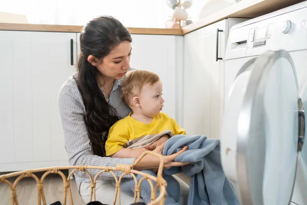 Beyaz tenli anne mutfakta bebek bebekle ev işi yapıyor. Güzel, genç bir anne, küçük oğlu evde oynarken çamaşır makinesine çamaşır koyuyor. Aile Hizmetleri.