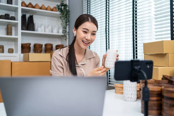 Asian beautiful woman sell vase product online live streaming at home. Young attractive girl use laptop computer shows goods to customer and present detail. Remote buying and purchase shopping concept