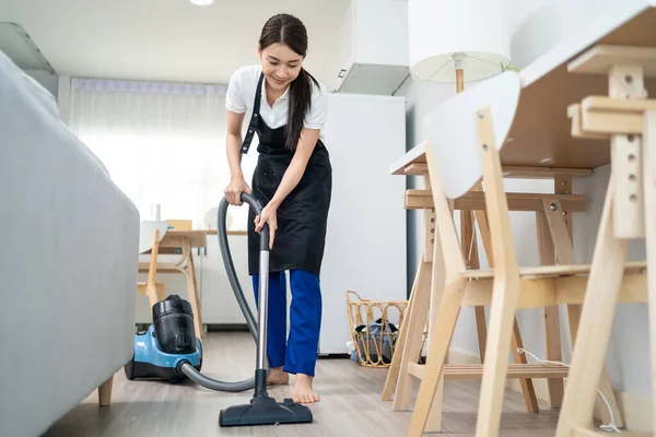 Asian cleaning service woman worker cleaning in living room at home. Beautiful young girl housekeeper cleaner wear apron and vacuuming messy dirty floor for housekeeping housework and chores in house.