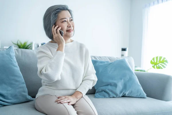 Asian senior old mature woman talking on mobile phone call with family. Attractive strong older female sit on sofa, spend time in living room at home, feel happy and enjoy communicate online at home.