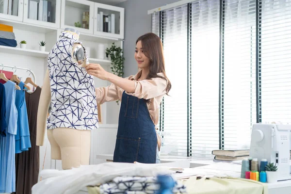 Asian beautiful tailor woman working on clothes in tailoring atelier. Attractive young girl fashion designer use tape measure new handmade suit on mannequin in workshop room. Fashion business concept.