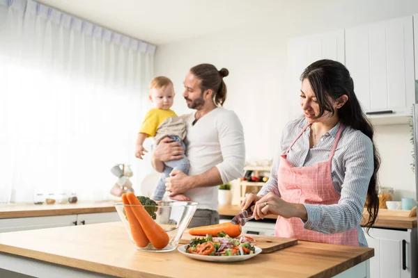 Caucasian beautiful parents cook food with baby boy toddler in kitchen. Happy family, Attractive young mother making healthy salad for lunch while husband father holding little kid son infant in house